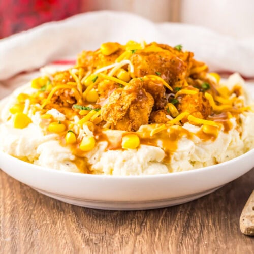 A white bowl filled with mashed potatoes, topped with fried chicken pieces, corn, shredded cheese, and brown gravy sits on a wooden table next to a wooden spoon.