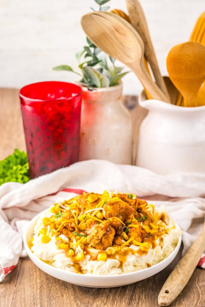 A plate of mashed potatoes topped with fried chicken, corn, gravy, and green onions, sits on a wooden table with a red glass, potted plants, and wooden utensils in the background.