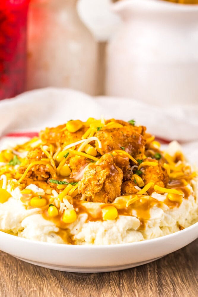 A plate of mashed potatoes topped with fried chicken pieces, corn kernels, shredded cheese, and brown gravy.