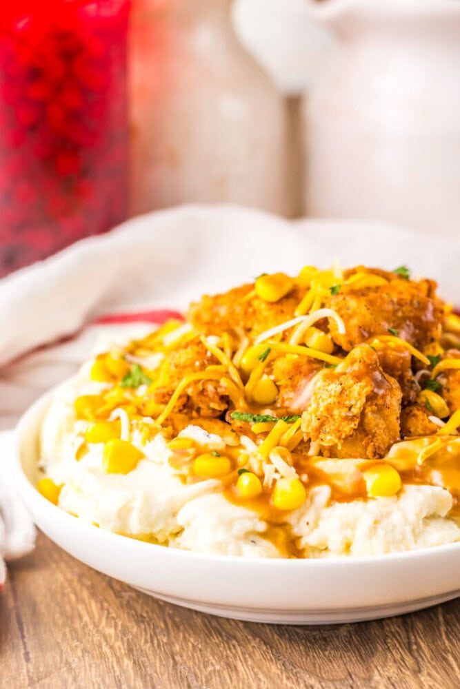 A bowl of mashed potatoes topped with fried chicken, corn, shredded cheese, and brown gravy sits on a wooden table.