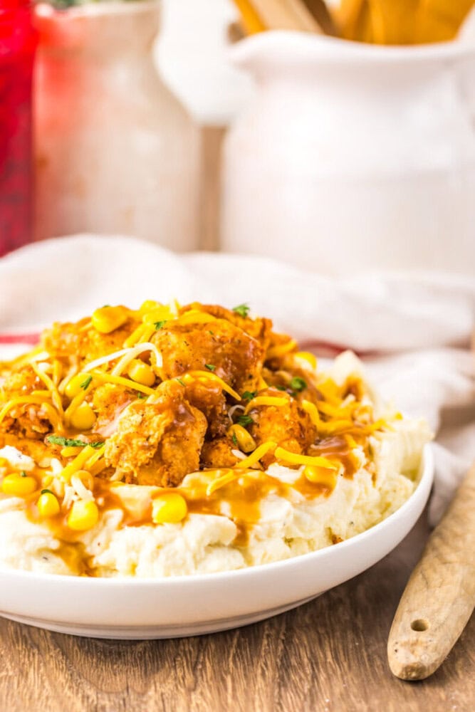 A bowl of mashed potatoes topped with fried chicken pieces, corn, shredded cheese, and brown gravy, set on a wooden table.