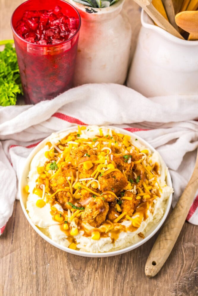 A bowl of mashed potatoes topped with fried chicken, gravy, cheese, and corn sits on a wooden table next to a red drink and kitchen utensils.