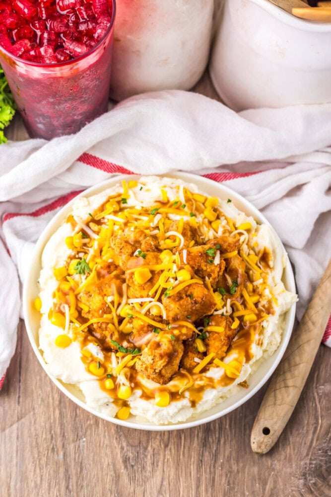 A bowl of mashed potatoes topped with fried chicken pieces, shredded cheese, corn, and brown gravy, with a drink and kitchen items in the background.