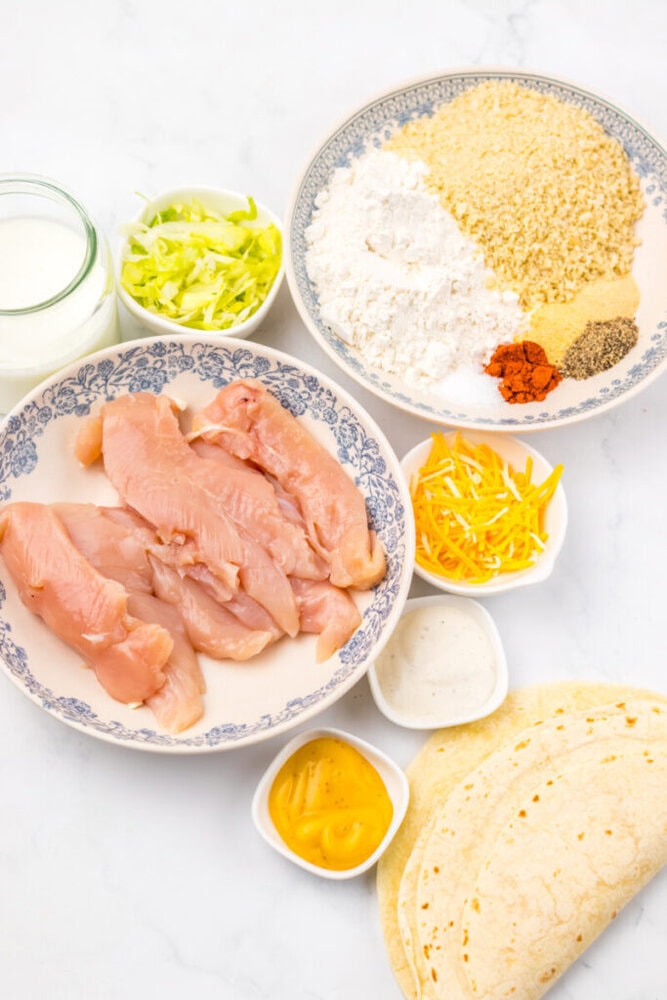 Raw chicken strips, shredded lettuce, flour, panko, spices, shredded cheese, tortillas, ranch, cheese sauce, and buttermilk displayed in bowls and plates on a white surface.