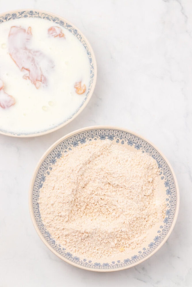 A bowl of breadcrumbs beside a bowl with pieces of meat soaking in milk, both on a white marble surface.