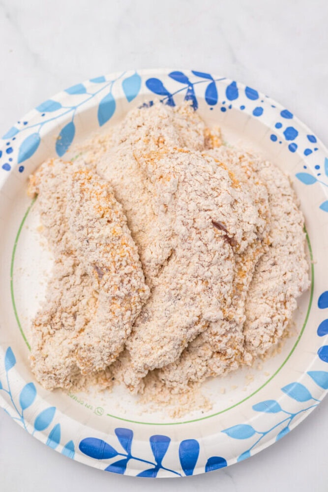 Raw chicken tenders coated in flour and seasonings are arranged on a paper plate with a blue leaf pattern.