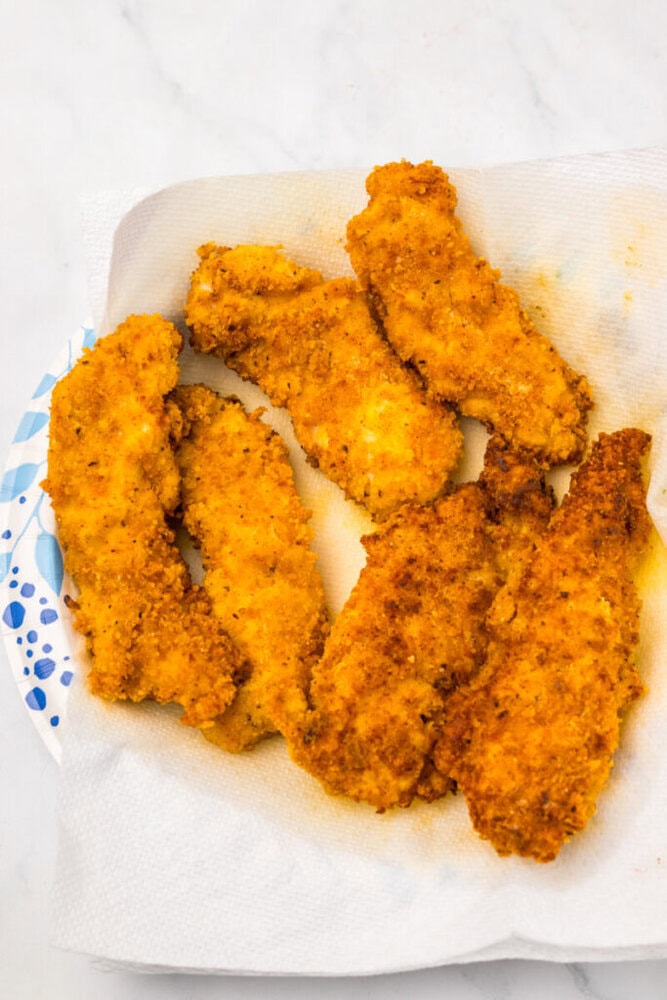 Six breaded and fried chicken tenders rest on a paper towel-lined plate, absorbing excess oil on a white marble surface.