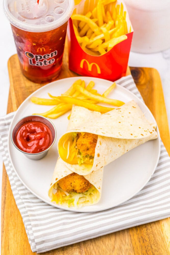 A plate with two McDonald's wraps filled with chicken and lettuce, a side of fries with ketchup, and a soft drink, all placed on a striped napkin and wooden tray.