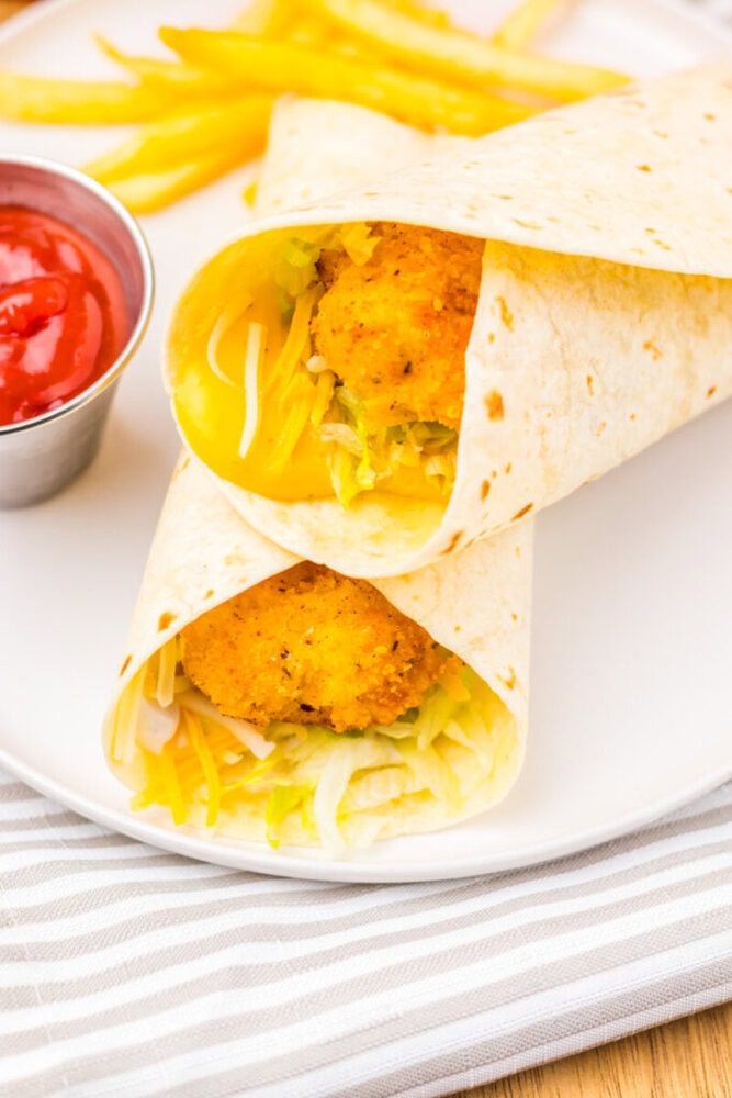 Two breaded chicken wraps with lettuce and shredded cheese on a white plate, served with fries and a cup of ketchup.