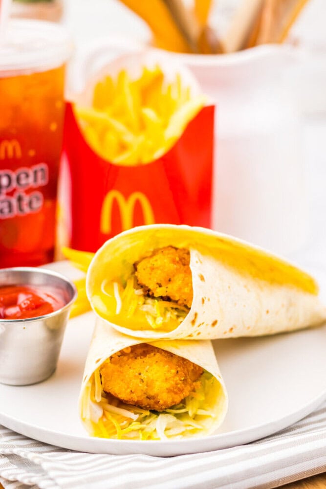 A McDonald's wrap with crispy chicken, shredded lettuce, and cheese, served with fries, ketchup, and a soft drink on a white plate.