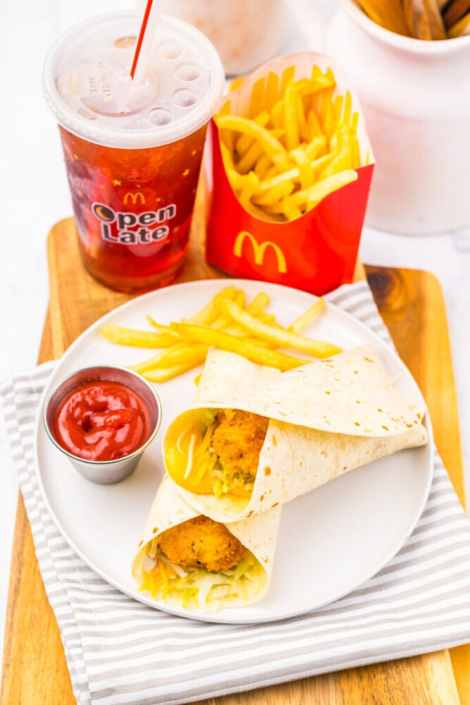 A McDonald's meal with a cup of soda, fries in a red container, a plate with two chicken wraps, extra fries, and a metal cup of ketchup on a striped napkin.