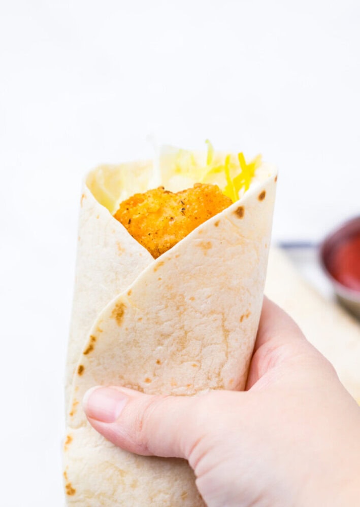 A hand holding a soft tortilla wrap filled with fried chicken, shredded cheese, and lettuce.