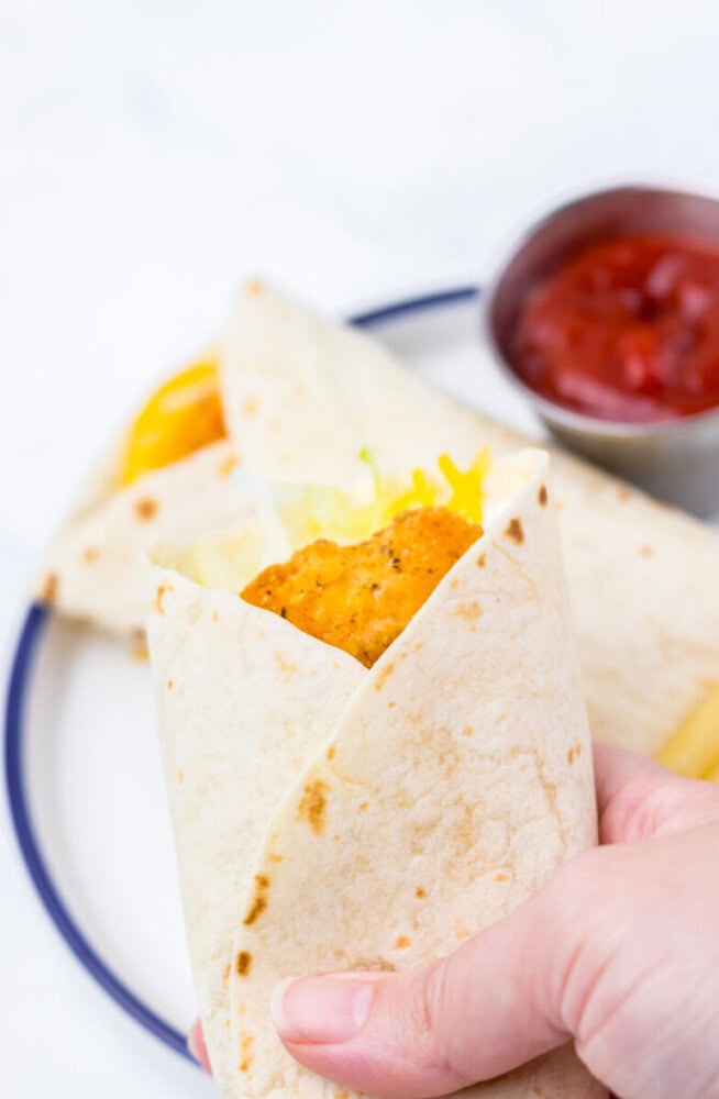 A hand holding a soft taco filled with crispy chicken, lettuce, and cheese, with another taco and a cup of salsa on a plate in the background.