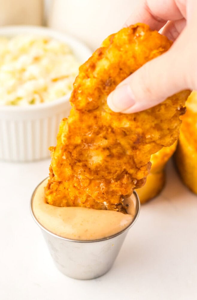 A hand dips a crispy fried chicken tender into a cup of creamy sauce, with a bowl of coleslaw and cornbread in the background.