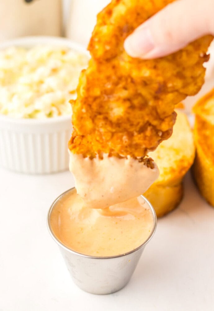 A hand dips a crispy chicken tender into a cup of creamy, orange dipping sauce. Coleslaw and slices of bread are visible in the background.