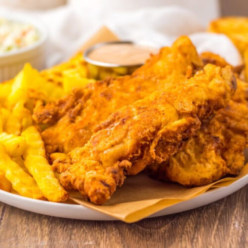 A plate of fried chicken tenders, crinkle-cut fries, a small cup of sauce, coleslaw, and toasted bread on a wooden table.