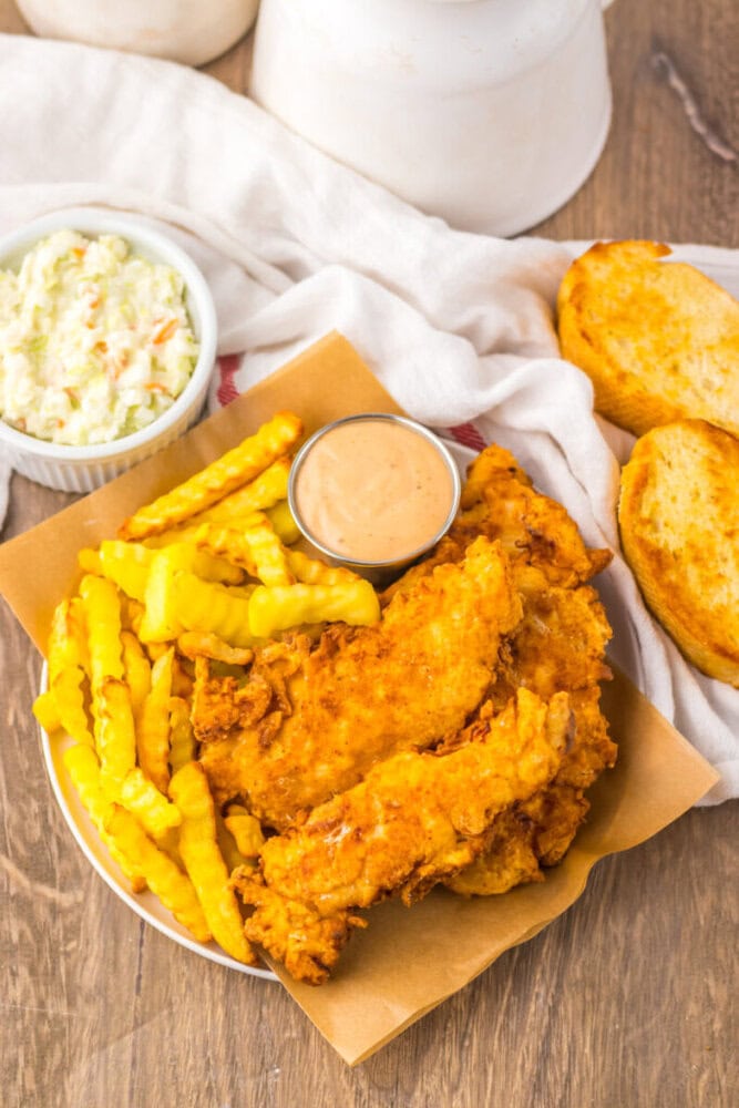 A plate of fried chicken tenders, crinkle-cut fries, a small cup of dipping sauce, coleslaw in a bowl, and two slices of toasted bread on a wooden table.