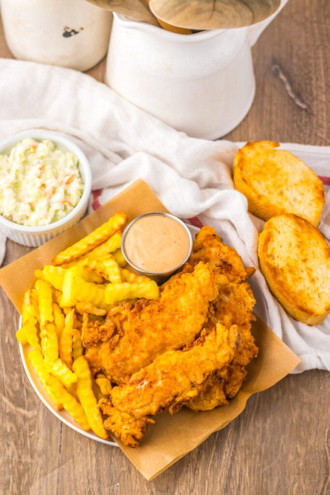 A plate with fried chicken tenders, crinkle-cut fries, a cup of dipping sauce, coleslaw in a bowl, and two slices of toasted bread on a wooden table.