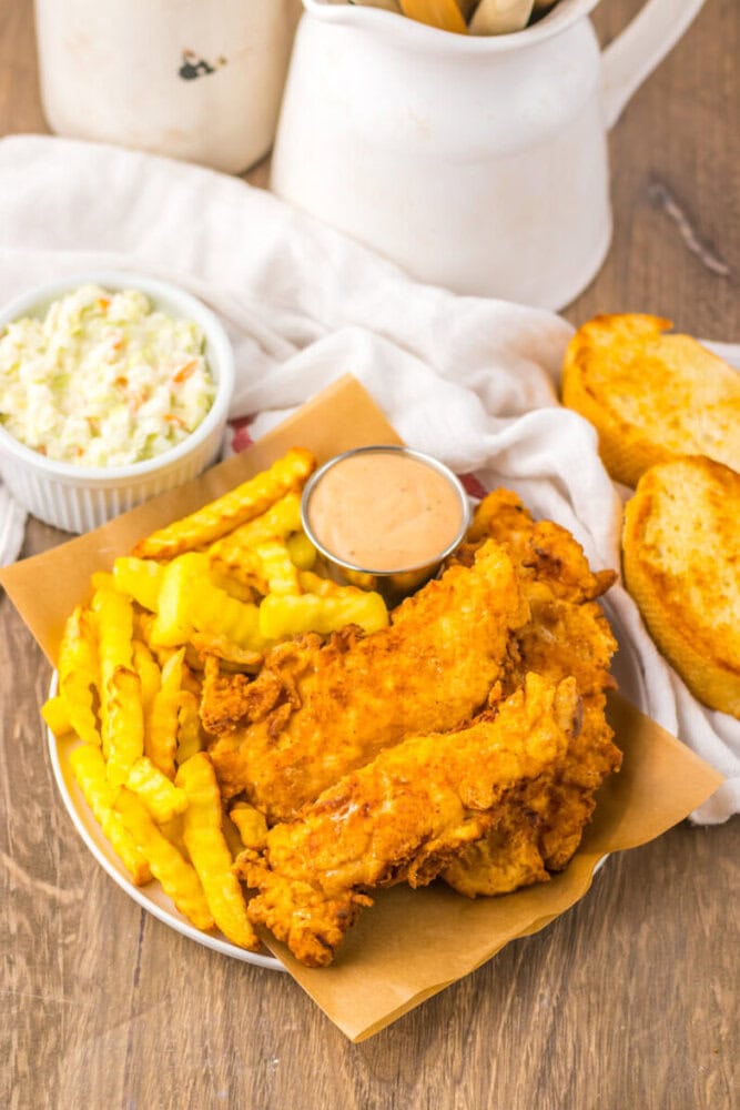 A plate of fried chicken tenders, crinkle-cut fries, dipping sauce, coleslaw, and two slices of toasted bread on a wooden table.