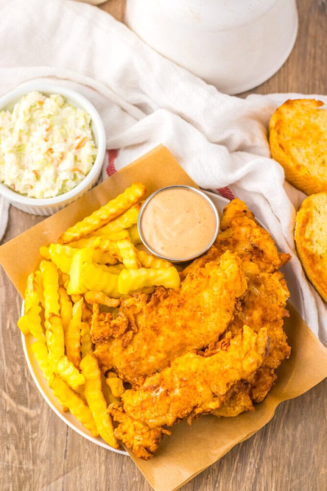 A plate with fried chicken tenders, crinkle-cut fries, dipping sauce, and toasted bread. A bowl of coleslaw is placed nearby on a wooden surface.