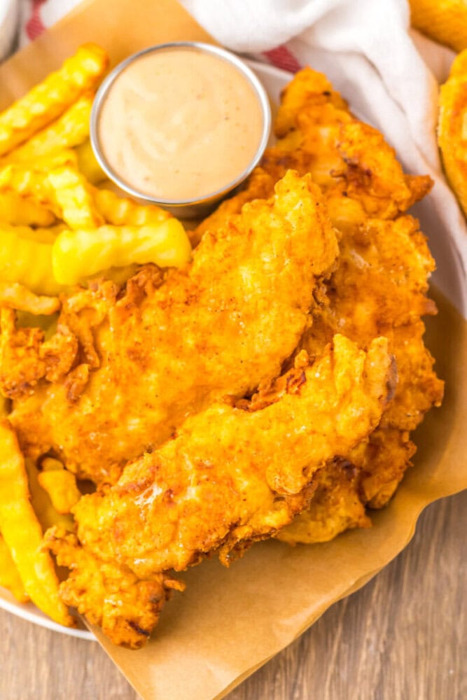 A plate of crispy fried chicken tenders with crinkle-cut French fries and a small cup of dipping sauce.