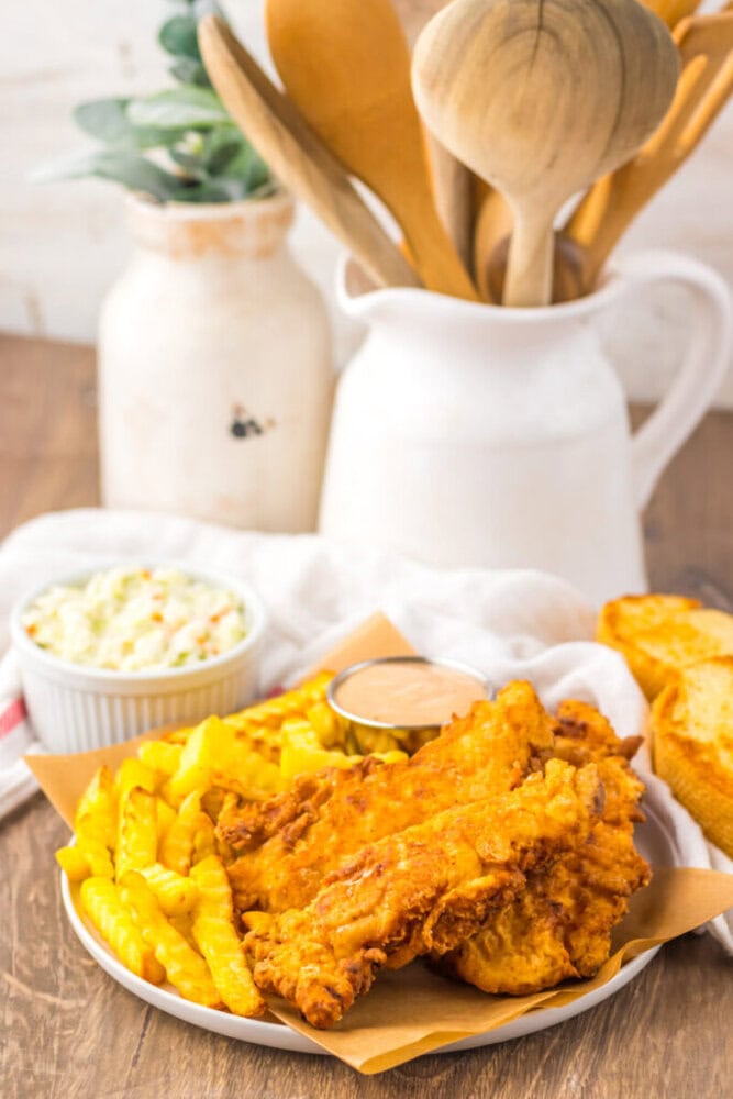 A plate with fried chicken tenders, crinkle-cut fries, a cup of dipping sauce, coleslaw, and a slice of toasted bread sits on a wooden table near ceramic pitchers with wooden utensils.