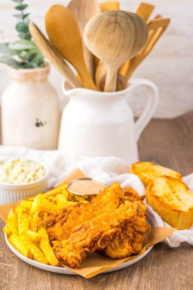 A plate of fried chicken tenders, crinkle-cut fries, garlic bread, dipping sauce, and coleslaw sits on a table with wooden utensils in a pitcher in the background.