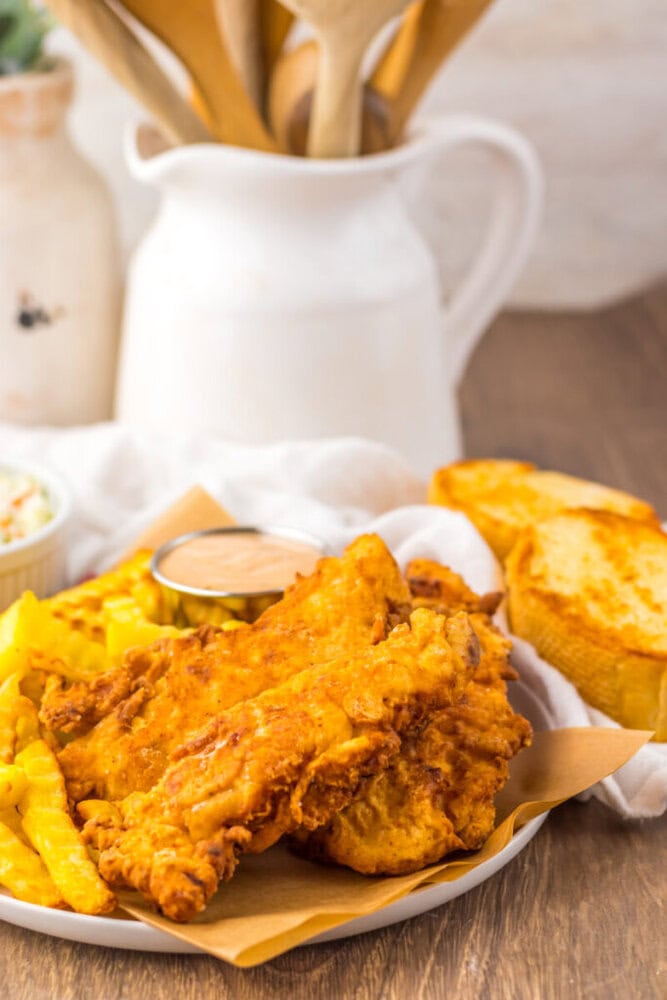 A plate of fried chicken tenders, crinkle-cut fries, dinner roll, and a small cup of dipping sauce, with a white pitcher and wooden utensils in the background.