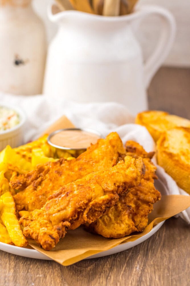 A plate of fried chicken tenders with crinkle-cut fries, toast, dipping sauce, and a small cup of coleslaw on a wooden table.