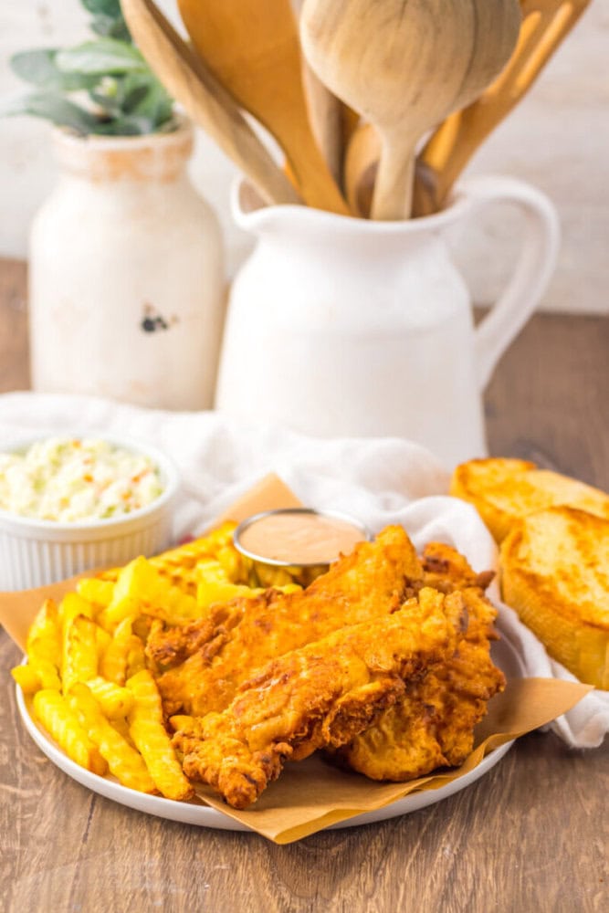 A plate of fried chicken tenders, crinkle-cut fries, a cup of dipping sauce, coleslaw, and a slice of toasted bread on a wooden table with kitchen utensils in the background.
