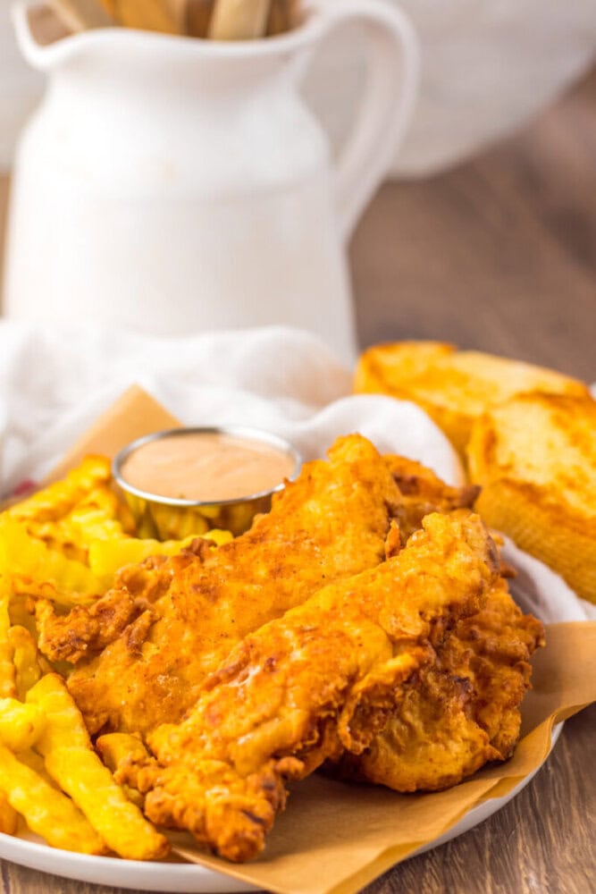 A plate of fried chicken tenders, crinkle-cut fries, a cup of dipping sauce, and slices of toasted bread on a table.