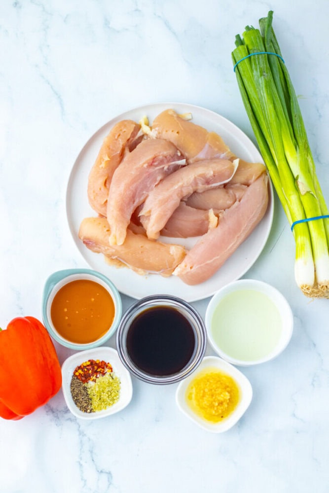Raw chicken strips on a plate, green onions, a red bell pepper, and small bowls of sauce, oil, minced garlic, and assorted spices on a white surface.