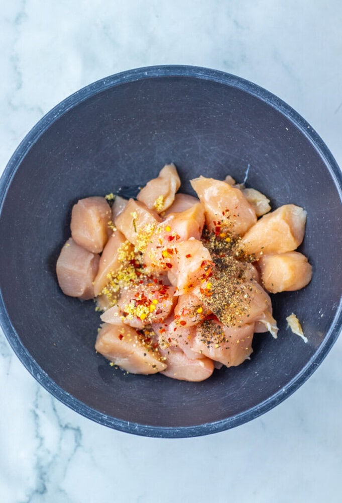 A black bowl with raw, cubed chicken breasts topped with various spices, including crushed red pepper and ground seasonings, on a white marble surface.