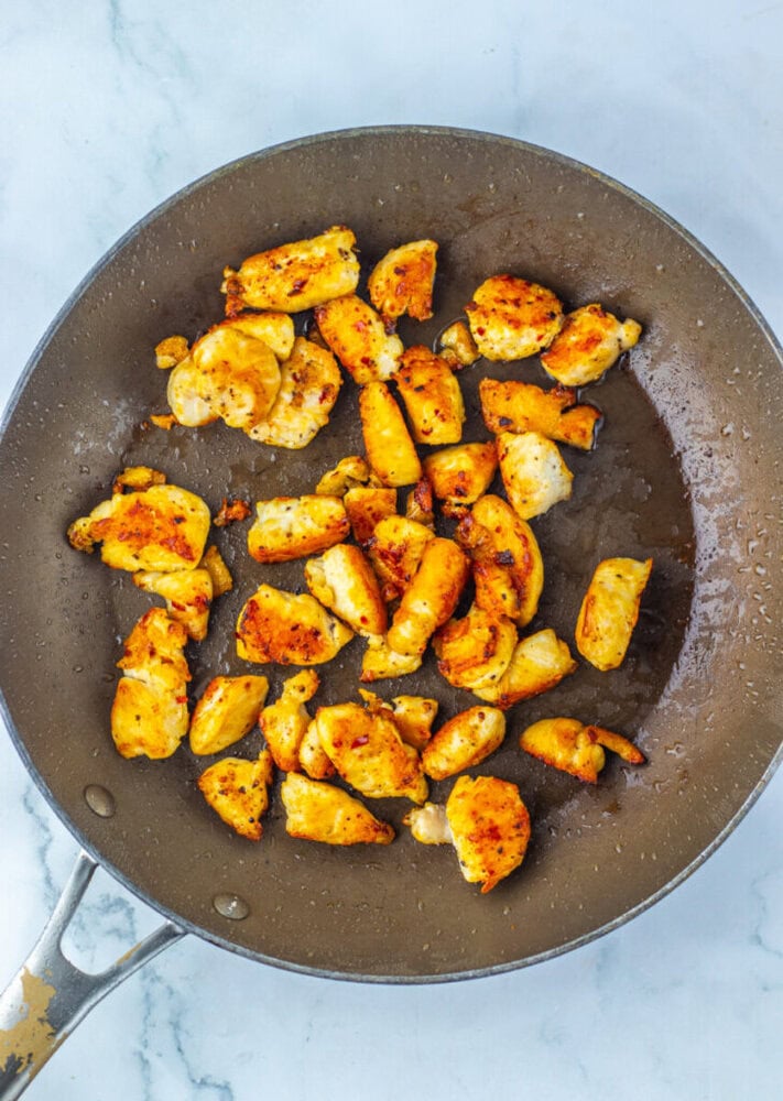 Cooked, browned chicken pieces are spread out in a nonstick skillet on a marble countertop.