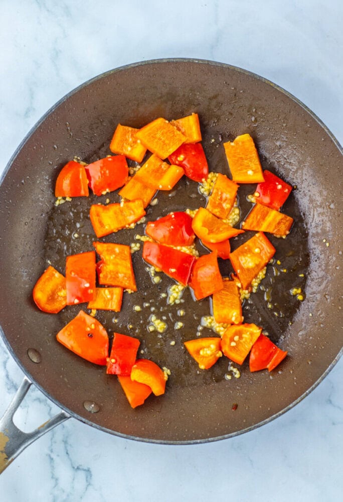 Chopped red and orange bell peppers being sautéed with minced garlic in a nonstick frying pan on a marble surface.