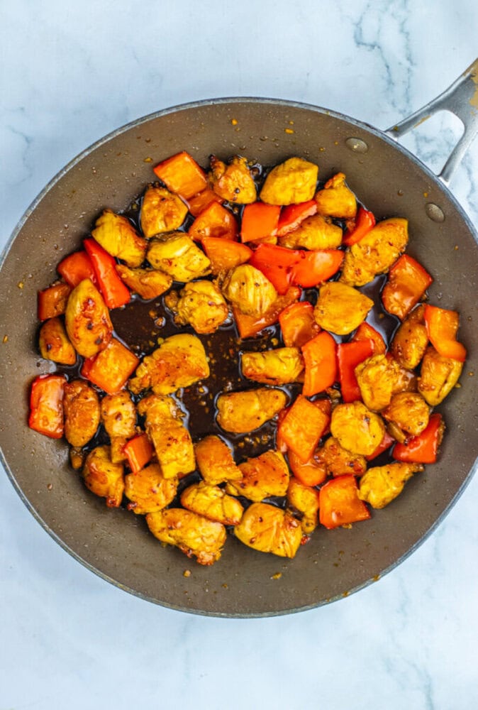 A frying pan containing cooked chicken pieces and red bell peppers in a brown sauce, set on a marble surface.