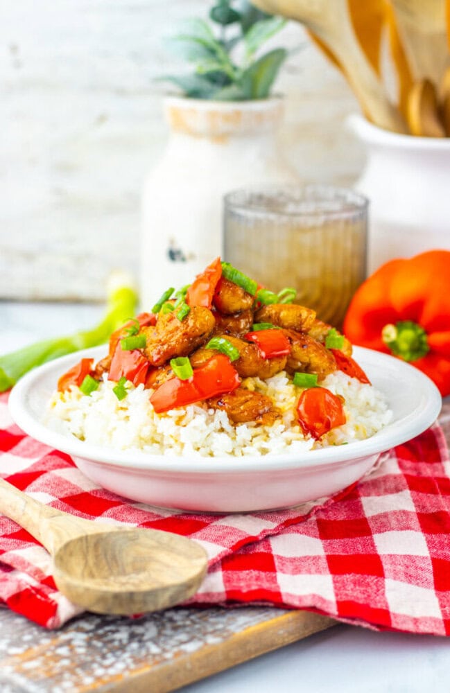 A white plate of rice topped with stir-fried chicken and red bell peppers, garnished with chopped green onions, sits on a red checkered napkin with a wooden spoon nearby.