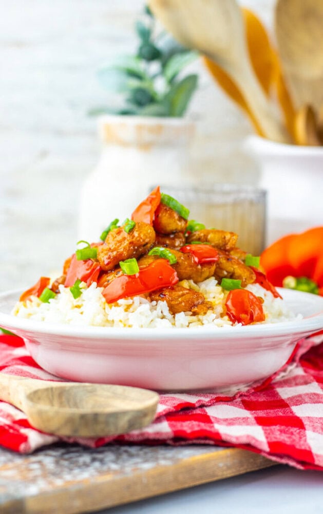 A white plate of rice topped with stir-fried chicken, red bell peppers, and green onions sits on a red checkered cloth with a wooden spoon nearby.