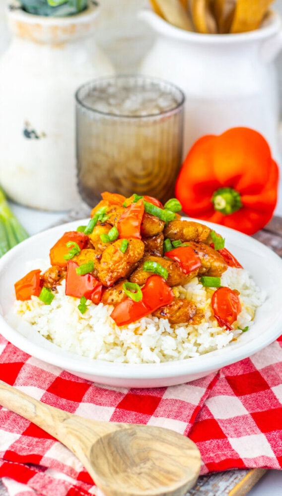 A plate of white rice topped with pieces of chicken, chopped red bell peppers, and green onions sits on a red checkered napkin with a wooden spoon beside it.