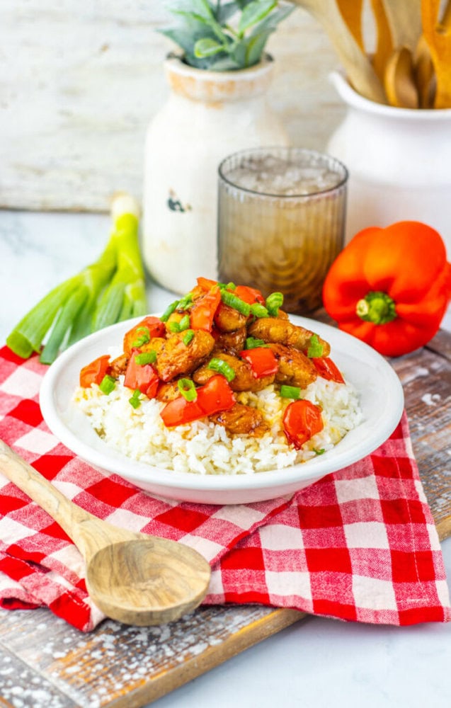 A plate of rice topped with cooked chicken, chopped tomatoes, and green onions sits on a red checkered cloth, with vegetables, a wooden spoon, and a drink in the background.