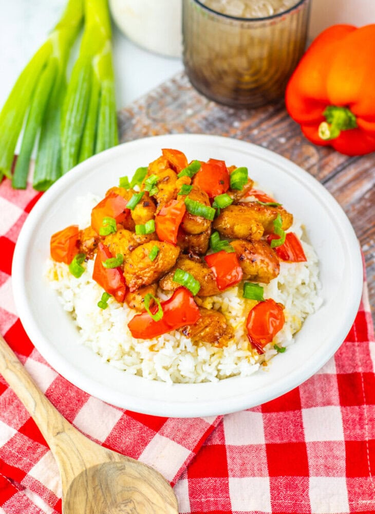 A plate of white rice topped with cooked chicken, chopped tomatoes, and green onions, placed on a red checkered cloth next to a wooden spoon, green onions, and a red bell pepper.
