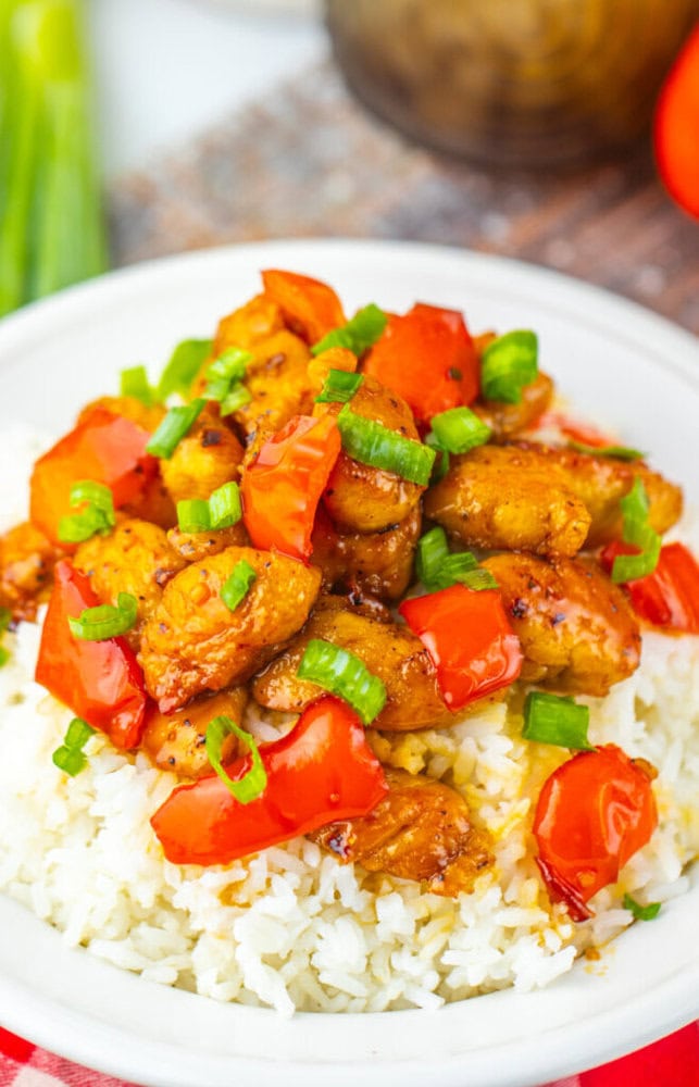 A white plate of white rice topped with pieces of cooked chicken, red bell peppers, and chopped green onions.