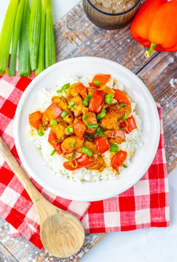 A white bowl of rice topped with sautéed chicken, red bell peppers, and green onions, placed on a red checkered napkin with a wooden spoon beside it.