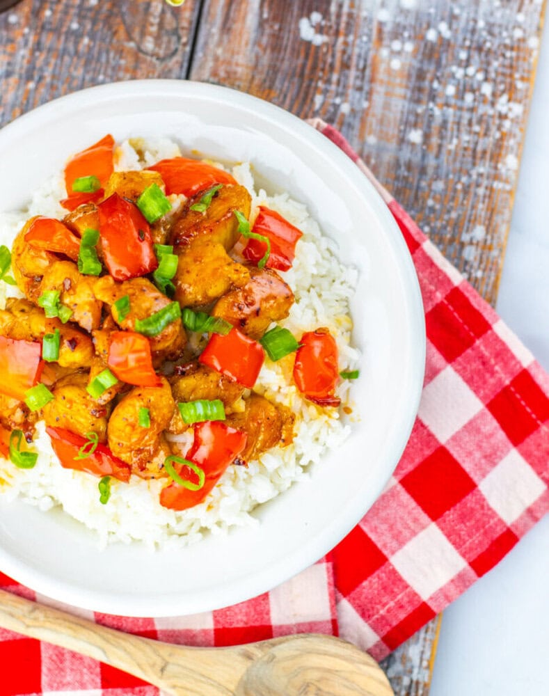 A white bowl filled with white rice, topped with shrimp, chopped tomatoes, and green onions, sits on a red and white checkered cloth on a wooden surface.