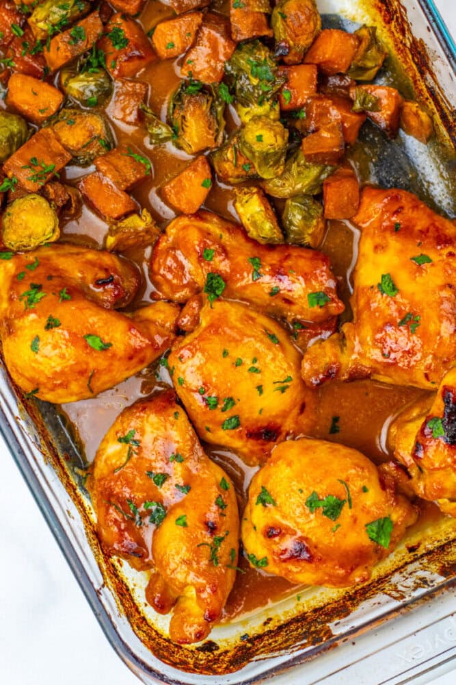 Baked chicken thighs with sauce in a glass dish, served alongside roasted sweet potatoes and brussels sprouts, garnished with chopped herbs.