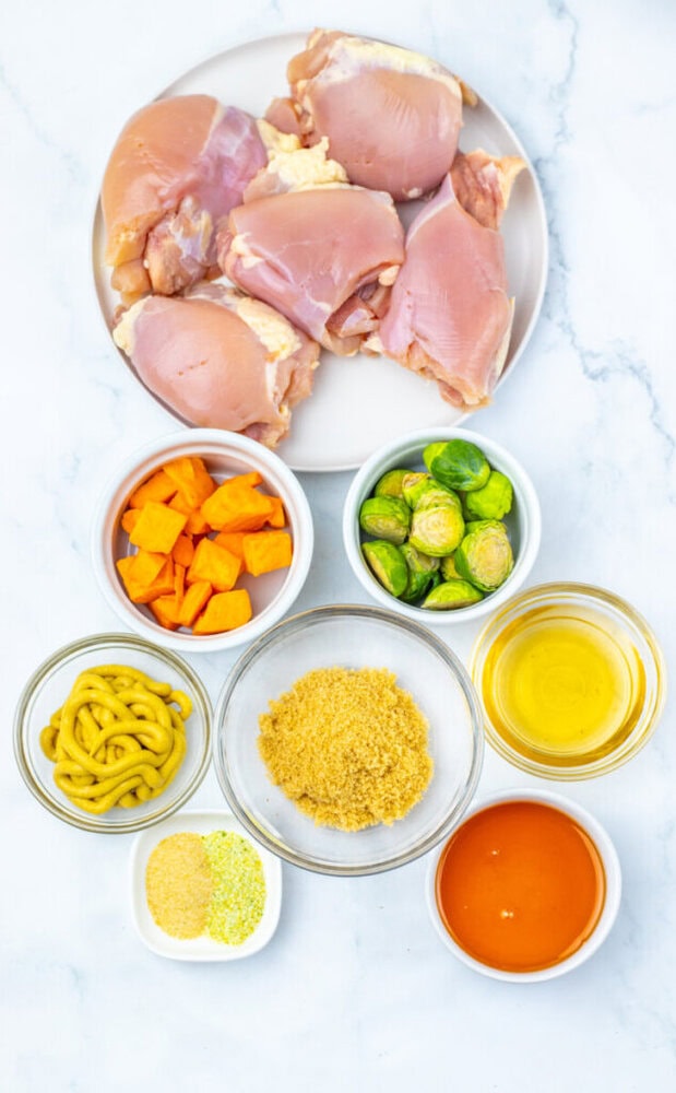 Plate of raw chicken thighs, bowls of chopped sweet potato, Brussels sprouts, mustard, brown sugar, apple cider vinegar, honey, and seasoning on a white surface.