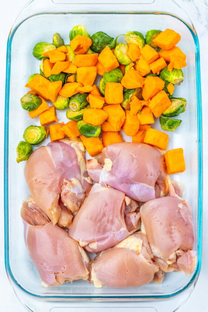 Raw chicken thighs, chopped sweet potatoes, and halved Brussels sprouts arranged in a glass baking dish, ready for cooking.