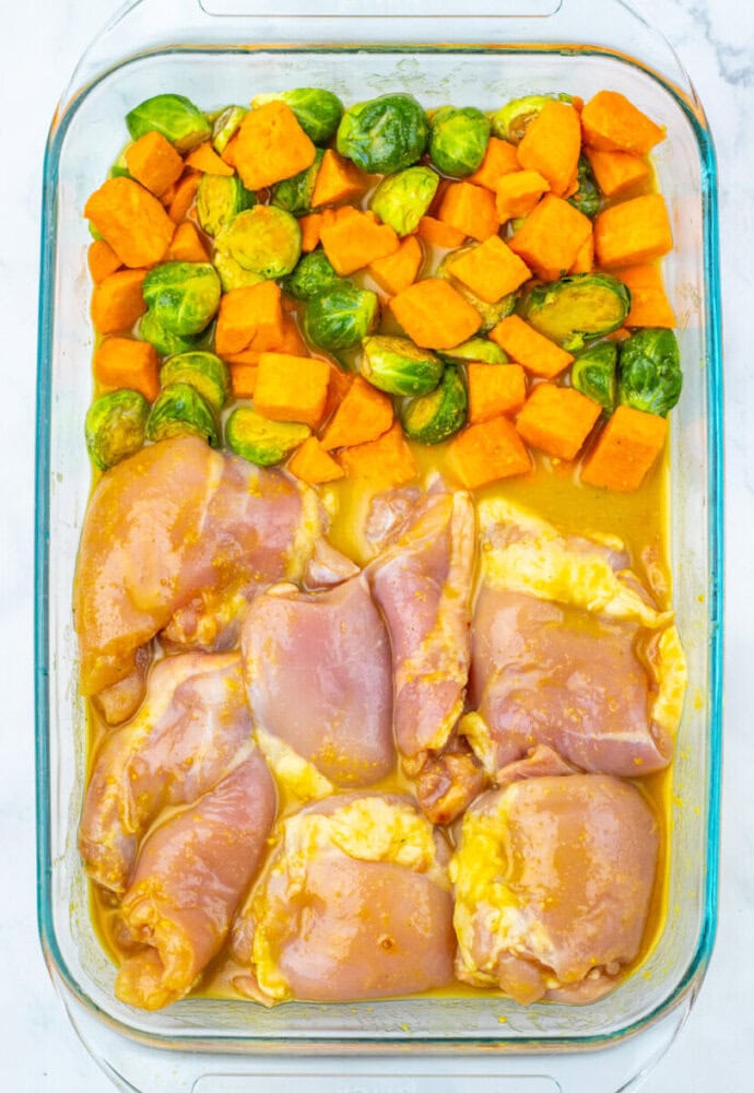 Uncooked marinated chicken thighs, sweet potato cubes, and Brussels sprouts arranged in sections in a glass baking dish.