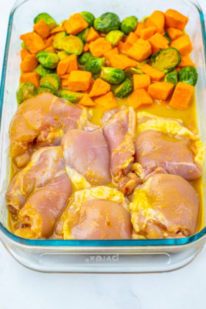 Glass baking dish with raw marinated chicken thighs on one side and chopped sweet potatoes and Brussels sprouts on the other, ready to be cooked.