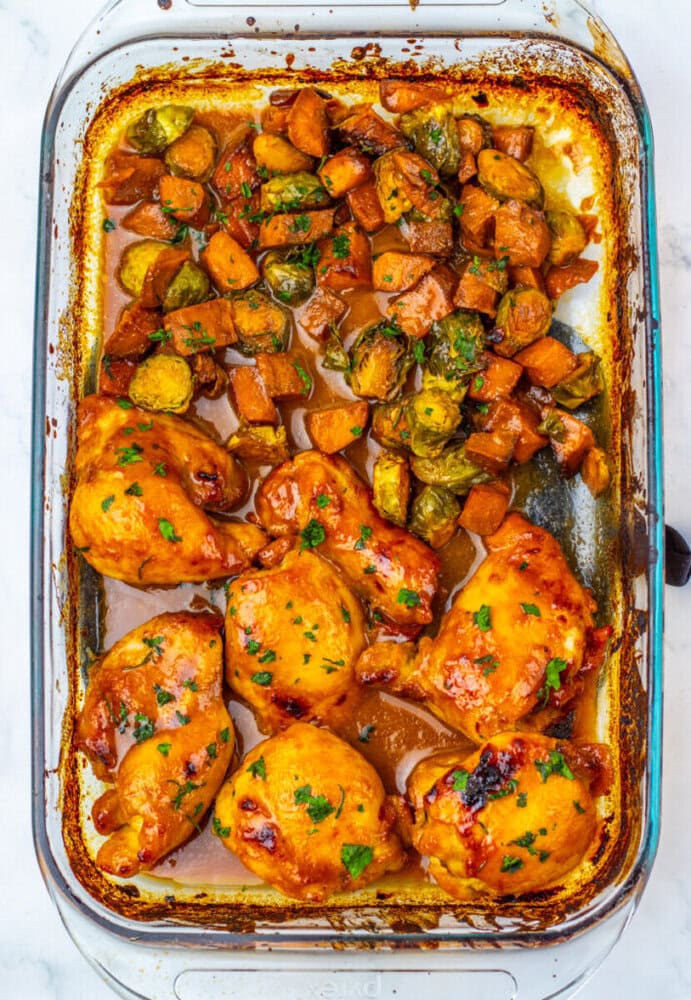 Baked chicken thighs and roasted vegetables, including Brussels sprouts and diced sweet potatoes, in a glass casserole dish with a glazed sauce.